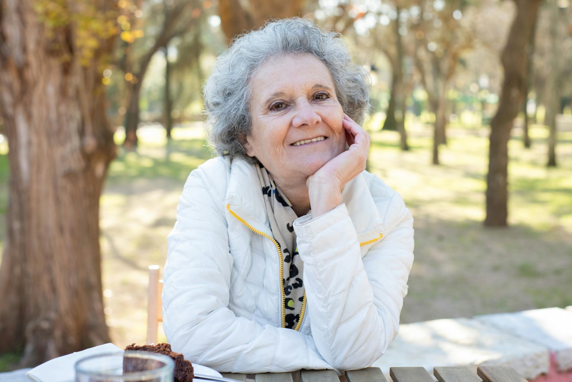 Donna anziana sorride serenamente all'obiettivo, seduta al tavolo da picnic in un bosco. L'attività all'aperto è ottima per l'invecchiamento attivo